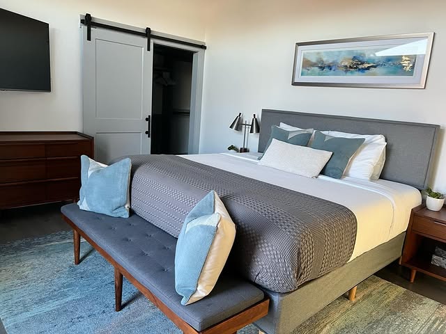 Impeccably cleaned modern bedroom with perfectly made bed, gray and blue accents, stylish barn door, wall-mounted TV, designer bench, and fresh linens - showcasing our thorough residential cleaning services in Raleigh
