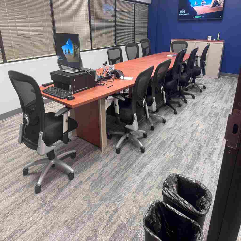 Clean conference room with long wooden table, ergonomic office chairs, blue accent wall, computer workstation, organized waste bins, and spotless carpeted floors - demonstrating our commercial cleaning expertise for Raleigh businesses